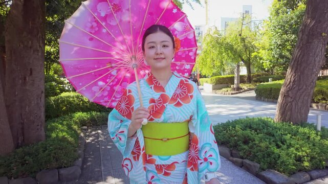 A young Japanese woman wearing a colorful kimono and holding a pink floral umbrella walks through Senso-ji temple in Tokyo, Japan, during summer.