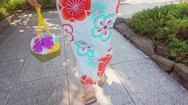 A woman walks along a stone path in Tokyo, Japan, wearing a traditional floral yukata and carrying a small bag on a sunny summer afternoon.