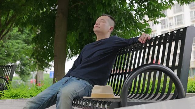 Man sits and rests on park bench. He sits on park bench and rests. He takes deep breath under the green trees in the park. Man tries to relax himself by taking deep breaths.