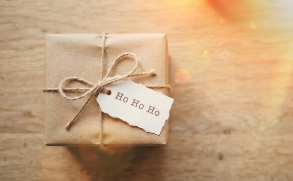 Wrapped Christmas gift box with brown craft paper and string bow with Ho Ho Ho tag on wooden table top view for holiday present concepts