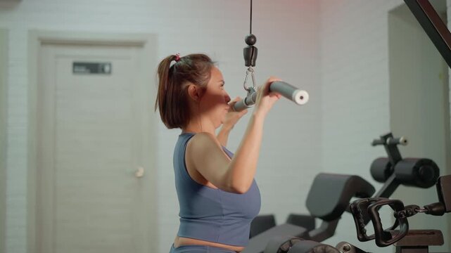 Woman in blue sports bra performs lat pulldown exercise using cable machine at home or commercial gym. White brick walls and closed beige door visible as background consistent lighting