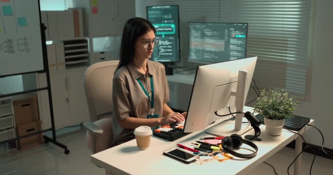 A young woman software engineer types on a mechanical keyboard at her desk, working through a block of code shown on the monitor in a bright modern technology office.

