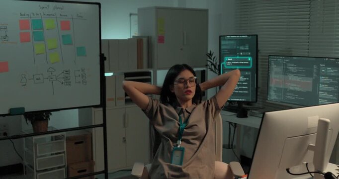  A young woman software developer leans back and stretches with her hands behind her neck, relaxing after finishing a coding task at her desk in a bright modern technology office
