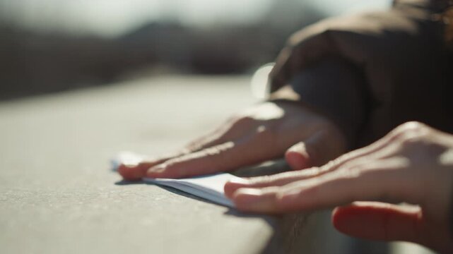 Close up view shows two human hands gently pressing and straightening edges of white papers placed upon smooth gray concrete. scene is softly lit by natural daylight creating warm shadows