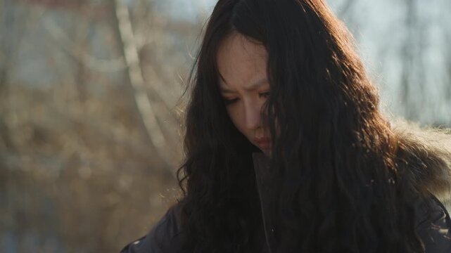 Close up shot captures upper body and head of young adult female with wavy black hair. Woman wears dark colored jacket standing in outdoor environment with blurred natural elements