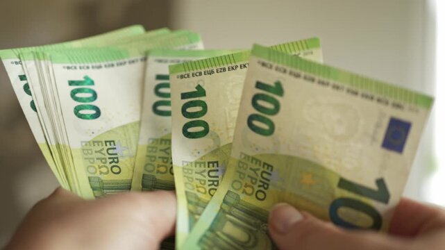 Hands display several 100 euro banknotes in a well-lit indoor environment, highlighting the intricate details and colors of the currency