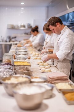 Chefs prepare bread in a professional kitchen. A team of chefs in white uniforms sprinkle flour on dough circles on a stainless steel counter;baked goods are being prepared, chefs are working together