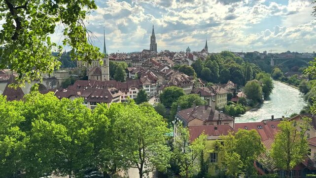 Historic Bern skyline appears beyond leafy trees and the Aare River outdoors, Elevated city view shows cathedral towers rooftops and Swiss architecture