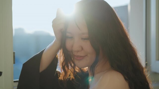 Close up portrait captures woman with wavy brown hair and dark eyes. Woman wears loose black off shoulder garment posing near open window that lets in bright sunlight creating soft lens