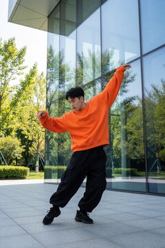 Young male dancer performs hip hop moves in urban city. Stylish guy wears bright orange sweatshirt, black cargo pants. Energetic breakdance performance by trendy man near modern glass building with