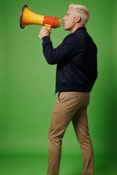 Young blonde man stands against green screen background. Holds yellow megaphone to mouth, shouts. Casual guy wearing dark jacket, khaki pants communicates loudly. Side view of male person using