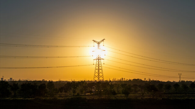 Silhouette of a high-voltage power transmission tower and lines against a vibrant sunset sky over a rural landscape.