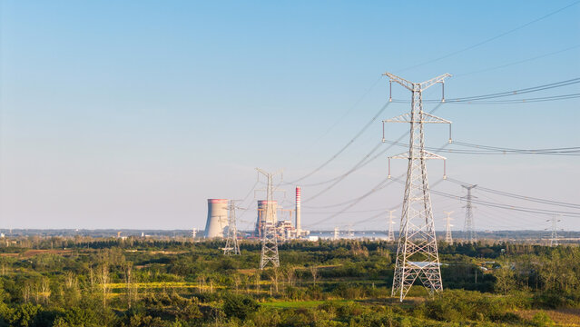 High-voltage power transmission towers and a thermal power plant stand in a rural vegetated landscape under a clear blue sky.