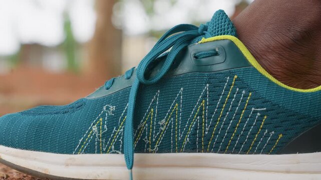 Woman teal sneaker is shown in detail as it rests near ground. shoe features intricate white and yellow stitching patterns on blue mesh fabric with matching laces tied neatly. Soft natural