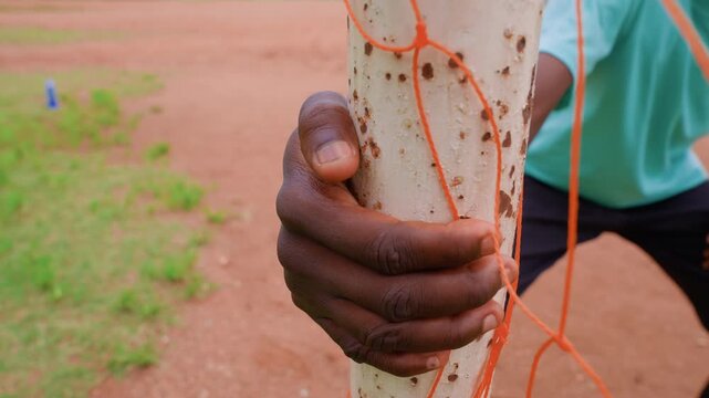 Shows someone tying orange string to vertical pole outdoors on reddish ground with green patches focusing on hand movements and interaction with objects