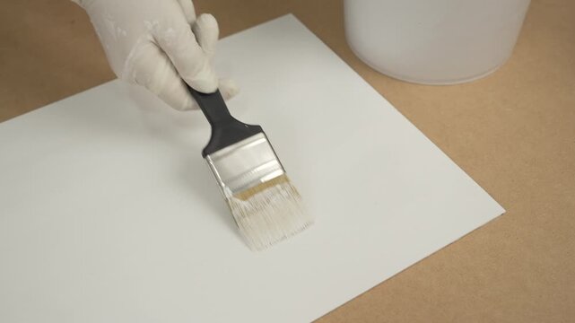 Gloved hand using a paintbrush to apply white paint or primer to a sheet of paper or board