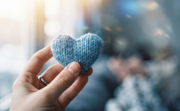 Hand holding a small knitted blue heart representing love and care in a cozy indoor setting