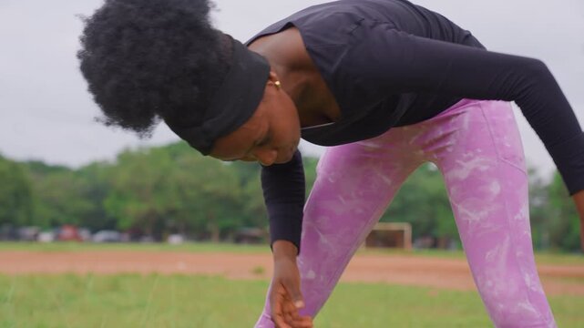 Young adult female stretches her legs and back on grassy field with trees. Woman wears black long sleeve top and pink patterned leggings bending forward to reach for ground