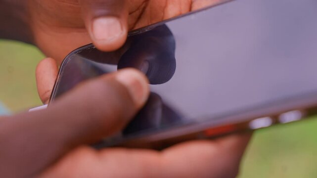 Close up shows two dark skinned fingers holding electronic gadget. One hand grips while another taps or swipes across its reflective surface suggesting active use of mobile device