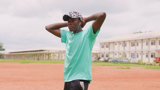 Young man performs dynamic poses and stretches on wide sports field. Man wears casual sportswear including aqua t shirt black shorts patterned cap and earphones. Buildings are visible