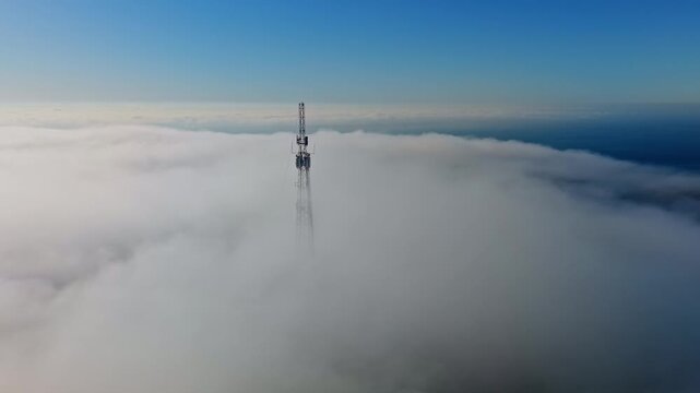 Aerial view of a telecommunications tower rising through dense coastal cloud and fog, with the ocean visible beyond under a clear blue sky.