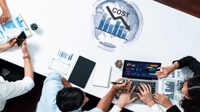 A diverse team is engaged in a meeting analyzing financial data and cost reduction strategies. They use charts and laptops to enhance productivity and collaboration. Raster