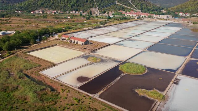 Drone video of the historic Ston salt pans features geometric evaporation pools in dynamic earth tones, rustic buildings, lush greenery, and scenic hills under sunlight.