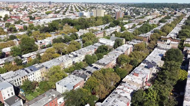 A drone glides forward and tilts down above tree lined Brooklyn row houses in New York City. The view changes from long residential streets to an overhead layout of rooftops gardens and intersections.