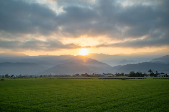 Morning sunlight breaks through clouds over lush green rice fields and hazy mountain ranges in Yuli, Taiwan.