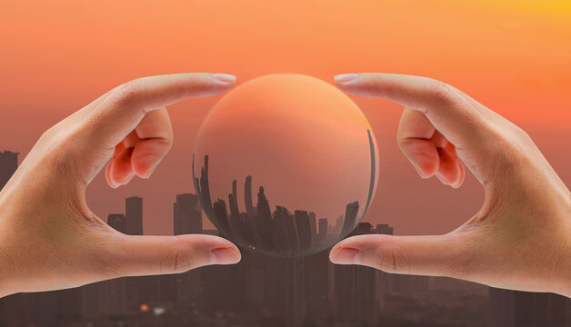 Human hands holding a glass sphere over a smoggy city skyline at orange sunset, illustrating air pollution, carbon capture technology and urban environmental protection for cleaner future living.