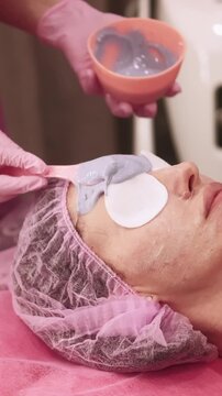 Vertical video. Skin cleansing. Detox procedure. Woman in beauty salon with cotton pads on eyes cosmetologist hands applying alginate mask with spatula on face.