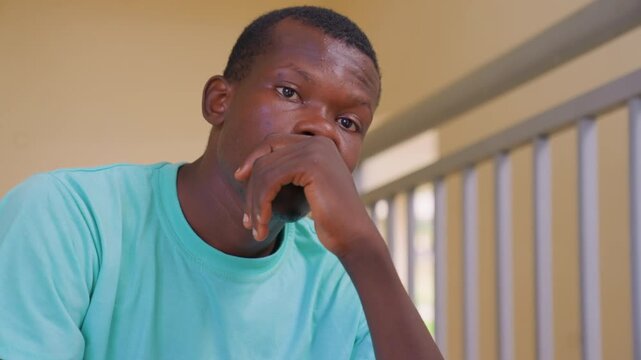 Young adult male rests thoughtfully at edge of outdoor porch. His posture suggests deep reflection or mild distress as man supports his head with one hand