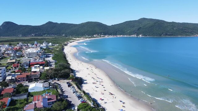 Drone aerial view of tourists relaxing on Praia dos Acores beach in Florianopolis, Brazil, featuring colorful umbrellas, white sand coastline, turquoise ocean waves, and green coastal mountains
