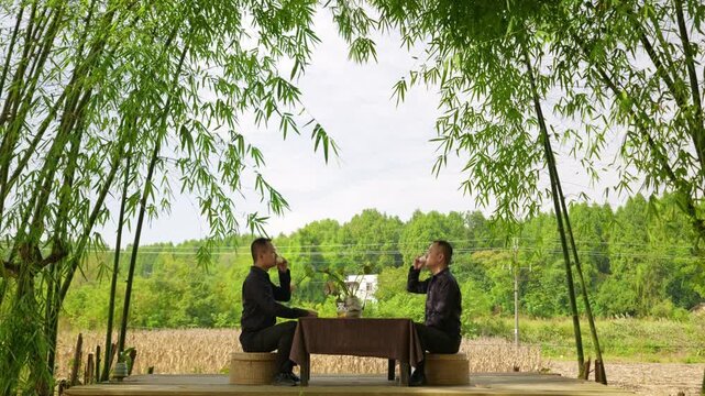 Two men sit facing each other and drink tea. Two men drink tea together in bamboo forest. (The scene is created in post production and actually features the same person.)