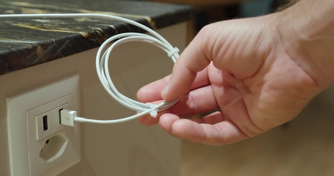Close-up of a person securing a coiled charging cable with a cable tie near a power outlet, representing cable organization, storage and household tidiness.