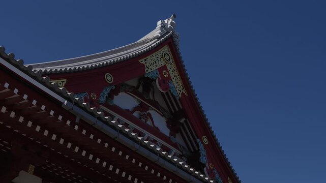 Vermilion gable end of a Japanese temple hall with gilded carvings, manji emblems and upturned tiled eaves under a clear sky, seen from below.