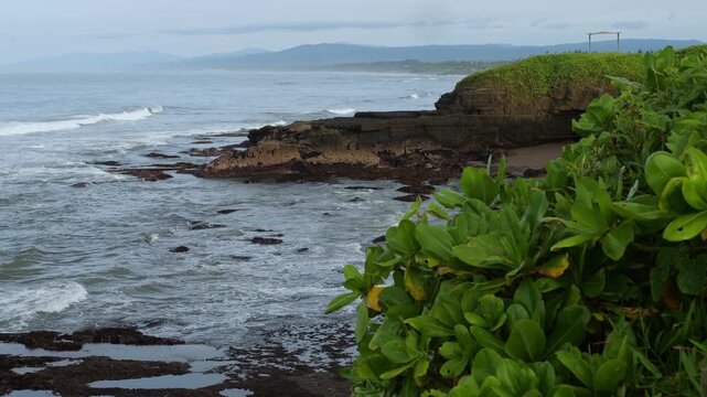 Secluded Bali Black Sand Coastline View With Basalt Rocks And Tidal Pools, Gentle Waves Lapping Empty Beach, Green Headland And Lush Foliage In Foreground, Drone Sweep Capturing Remote Bay