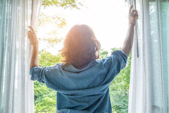 The woman was at the window in the bedroom. She opened the curtains on the window. In the morning and she looks at the view of mountains and trees at sunrise.