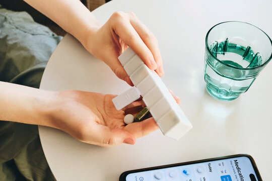 Woman taking medication from weekly pill organizer with glass of water and smartphone