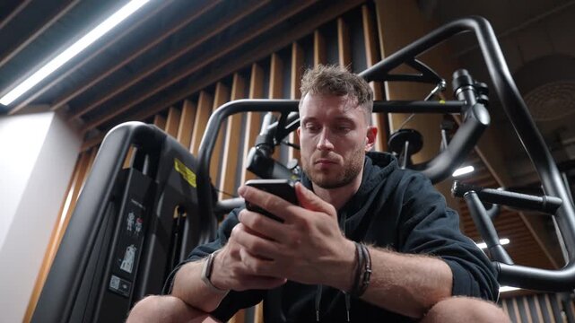 Low angle close-up of an athletic man sitting in a modern gym and scrolling on his smartphone instead of exercising in slow motion.