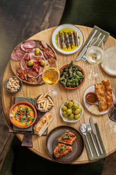 Top-down view of a round wooden table filled with Spanish tapas including charcuterie, olives, padron peppers, anchovies, focaccia, and white wine.