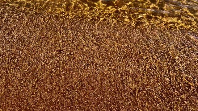 Clear shallow water flows continuously along sandy beach edge. Sunlight illuminates tiny golden ripples as ambient outdoor current recedes smoothly over fine pebbles under warm radiant beams.
