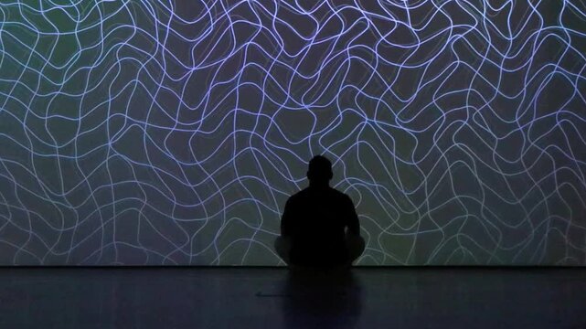 Silhouette of man meditating in front of glowing light line pattern wall in dark room