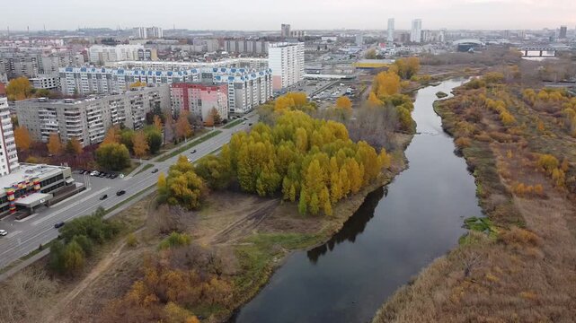 Aerial view of river flowing through autumn cityscape. Buildings line the river's edge with vibrant yellow foliage. Roads run parallel to the water, bustling with daily traffic