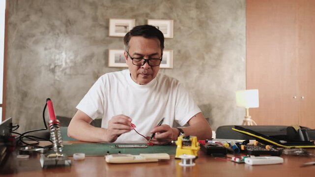 Mature Asian man checking soldering circuit board job with multimeter, cheerful, arms crossed with job achievement pride, home electronics appliance repair successful, DIY fix tools, hobby workshop.