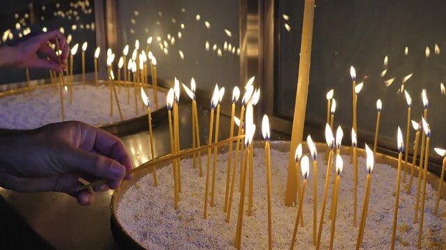 Hands performing a religious ritual, lighting a thin candle among many flickering flames in a container of sand in an Orthodox church in Greece
