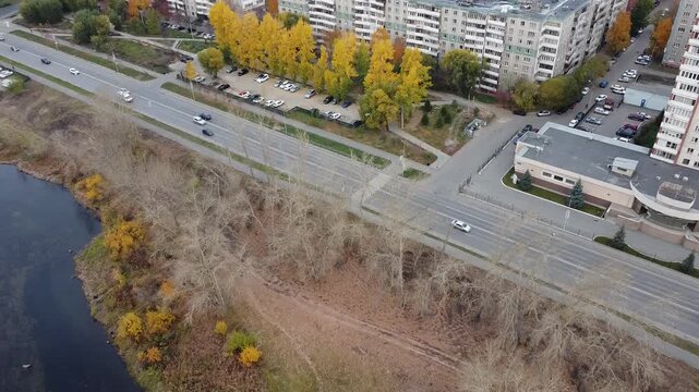 Aerial view of urban riverbank with autumn trees. Road runs parallel to water, cars move steadily. Buildings line the streets, showing city life. Fall colors contrast with dry earth and water