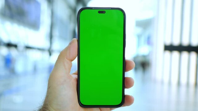 Hand holds phone with green screen. Background shows blurred airport terminal. Three keyframes show same scene. Focus remains on phone throughout