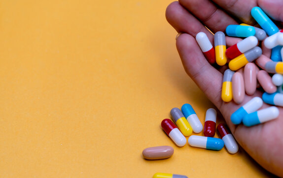 Hand holding assorted colorful capsules and oblong tablets over a yellow surface, with loose pills nearby, associated with antibiotic overuse, antimicrobial resistance and superbug awareness.