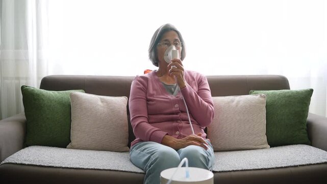 Older woman using a nebulizer for respiratory treatment while seated in her living room at home